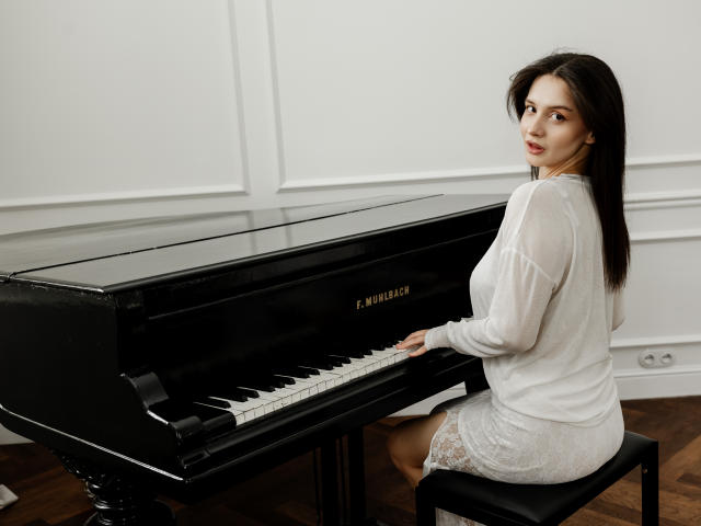 JeanieCat with tits visible in white long-sleeve top and lace dress, sitting on black stool playing black Steinway piano, wooden floor, looking over shoulder, dark brown hair.