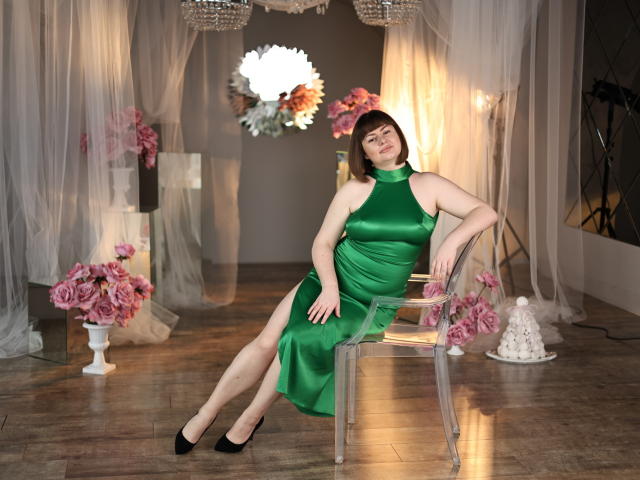 ZannaDreil, sitting on a transparent chair, wearing a green satin dress with a high neckline, black heels, and short brown hair, legs crossed, with a confident, relaxed expression.