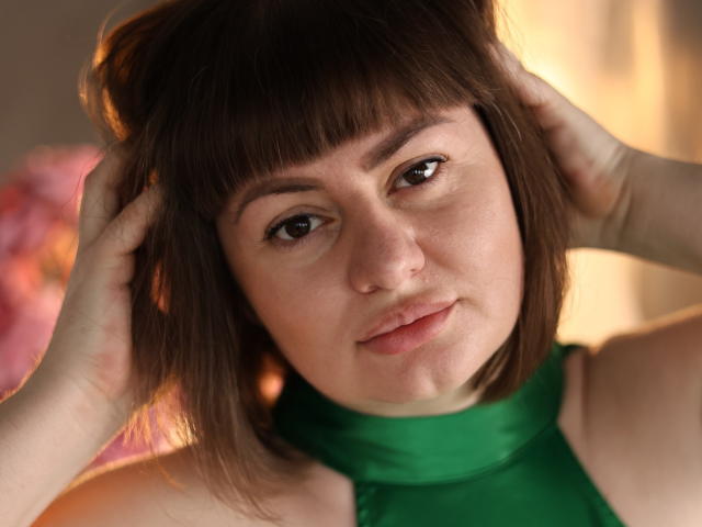 ZannaDreil with brown bob and green halter top, hands in her hair, looking at the camera with a slight, confident smile, soft light highlighting her fair skin and freckles.