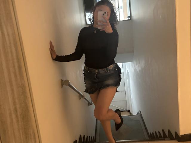 SofiaPassionne with curly hair, black long-sleeve top, denim mini-skirt, black high heels, standing in a narrow hallway, holding phone, showing off her ass and thighs.