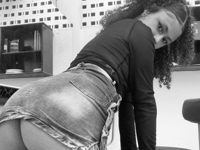 SofiaPassionne with curly hair, black long-sleeve top, and denim skirt, bending over, showing her ass, looking back with a seductive gaze in a black-and-white photo.