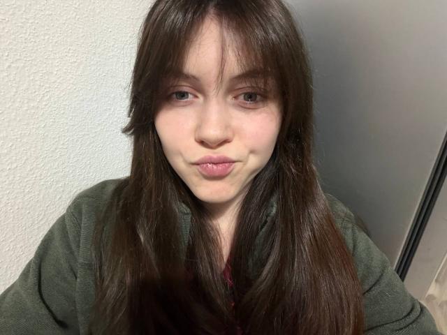 BellPrisco has long, straight brown hair with bangs covering her forehead. She's making a slightly pouty, closed-lip expression. Her skin is pale with a few small blemishes. She wears a green, long-sleeved shirt. Her eyes are brown and slightly squinted. The texture of her hair is smooth, and her eyebrows are thin. The lighting is even, highlighting her facial features. The background is a plain, textured white wall. The image is a close-up selfie.