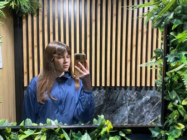 BellPrisco with long brown hair stands in a green, leafy environment, holding a phone in a reflective, marble-framed mirror. She wears a blue raincoat and black gloves. The mirror's reflection shows vertical wooden slats behind her. The image has a natural, realistic style with a focus on texture and contrast between the wooden slats and greenery. BellPrisco's expression is neutral.