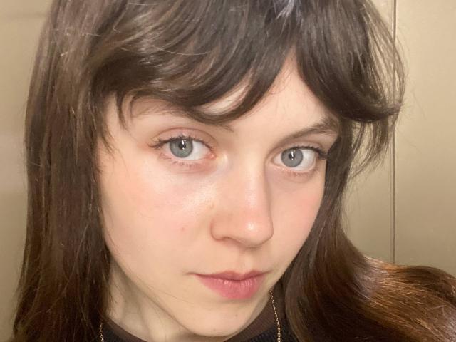 BellPrisco's face, pale skin, blue eyes, brown wavy hair with bangs, slight pinkish blush, subtle smile, wearing a black top, looking directly at camera.