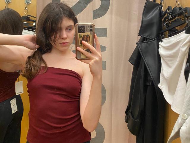 BellPrisco, a young woman with fair skin and brown hair, takes a mirror selfie in a clothing store. She's wearing a strapless, dark red top that clings to her slender frame. Her right arm is raised, hand touching her hair. She holds a phone with a brown case featuring a decorative design. Behind her, clothes hang on black hangers: a black leather jacket and a white shirt. The mirror reflects her side profile, and the background has a beige curtain with partial text. The image is a casual, realistic selfie with natural light.