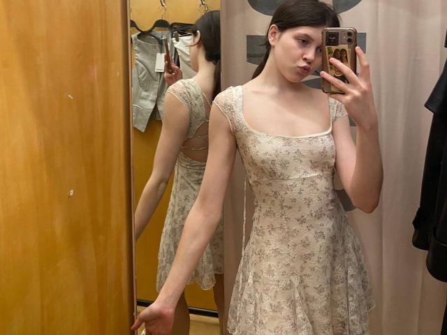 BellPrisco stands in a dressing room, taking a mirror selfie. She wears a light, floral, lace dress with a square neckline and short sleeves. Her dark brown hair is pulled back. She holds a gold smartphone in her right hand. The reflection in the mirror shows her back, highlighting the dress's delicate pattern and her slender build. Her left hand rests on a wooden door. The lighting is even, emphasizing her fair skin and subtle makeup. The image has a casual, everyday style.