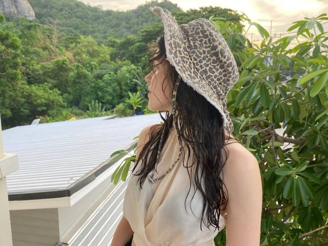 BellPrisco stands on a balcony, facing left. She has wet, dark hair and wears a leopard-print sun hat. Her beige, sleeveless top is slightly wrinkled. She has glasses and small earrings. Green foliage and a mountain are in the background. The balcony has a white, ribbed roof. Light filters from the right. Her skin is light. The image has a natural, relaxed feel.