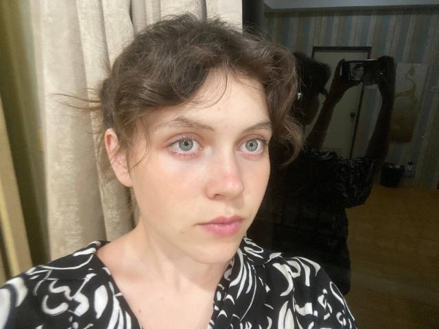 Close-up of BellPrisco with light skin, blue eyes, and wavy brown hair. She wears a black and white patterned shirt, looking slightly to the right. Her eyebrows are natural, and she has a subtle pink lip color. The background shows a reflection in a mirror and a curtain. The mirror shows a black bag and a camera. The composition is centered on BellPrisco's face, with soft, natural lighting highlighting her facial features. The texture of her shirt is smooth, contrasting with the wavy texture of her hair. Her expression is neutral, with slightly parted lips.