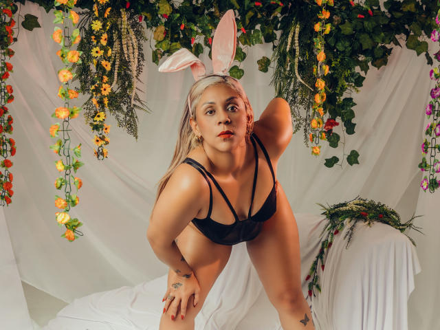 Viele Hawkey-Figuren mit rosa Hasenohren, schwarzem BH und Unterhose, die nach vorne gebeugt sind und ihren Hintern zeigen, mit Tattoos an Hand und Oberschenkel, blonden Haaren, vor einem weißen, drapierten Hintergrund, mit floralen Dekorationen..