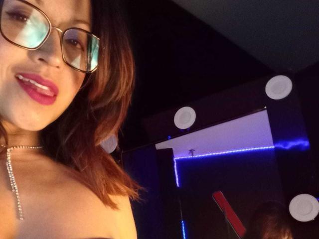 AngelinaBlanche with glasses, pink lipstick, and wavy brown hair, showing her bare shoulders and a silver necklace, smiling. Her tits are partially visible. Dark room, blue lights.