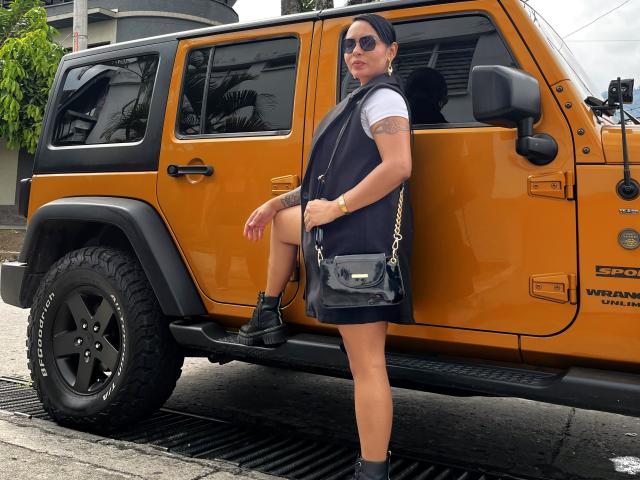 JessicaFiore stands in front of a yellow Jeep, wearing a black sleeveless vest, white shirt, black shorts, black boots, and a black crossbody purse, with one leg raised.