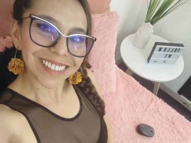VickyVegas has a light brown complexion, wearing blue-tinted glasses, and a sheer black top. She has a braid, yellow flower earrings, and a wide smile with visible teeth. The background includes a pink fuzzy pillow, a white circular table with a white vase holding green plants, and a sign that reads "WOLI" with a small "IN". A gray wireless mouse is on the table. The image is a selfie with a casual, friendly atmosphere.