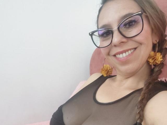 VickyVegas has light brown hair in a braid, wearing black, sheer, sleeveless top that reveals her small breasts. She has a fair skin tone, large purple eyeshadow, black-framed glasses, and is smiling with visible teeth. She has yellow flower earrings and is seated in a pink chair. The image is a close-up, focusing on her upper body and face. Her glasses have a slight reflection.