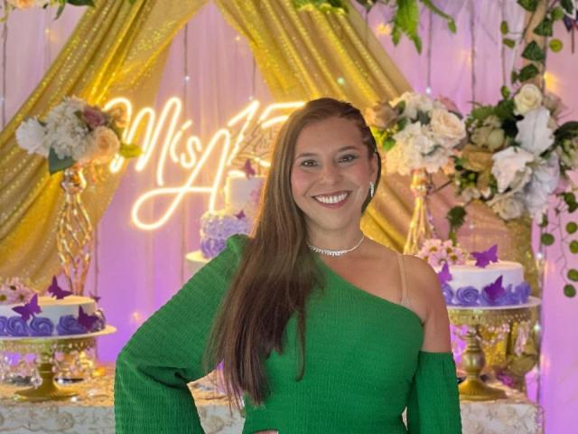 VickyVegas stands in front of a decorated backdrop with a neon "MIS A" sign. She wears a vibrant green, off-shoulder knit dress with long sleeves, and a sparkling choker. Her long, straight brown hair falls over her right shoulder. She smiles broadly, showing white teeth. The background includes floral arrangements, gold drapes, and purple and white cakes with purple butterflies. The image is brightly lit with warm, yellow-green lighting.