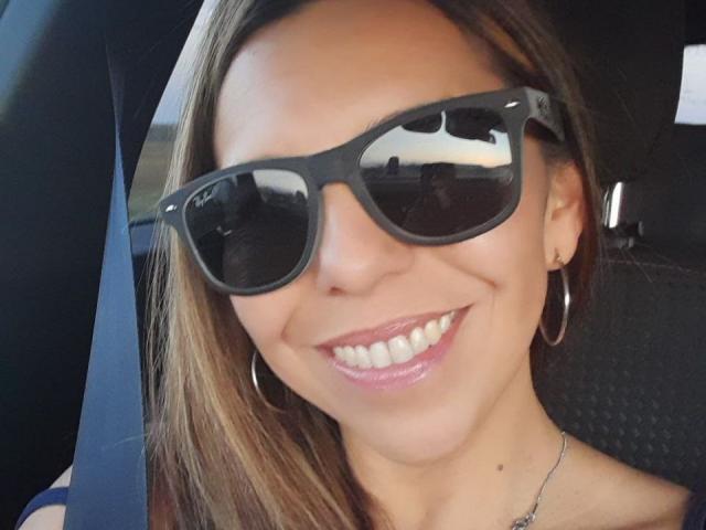 VickyVegas with shiny black sunglasses reflecting a car's interior, smiling with pink glossy lips, hoop earrings, and a seatbelt over her shoulder. She has light brown hair.