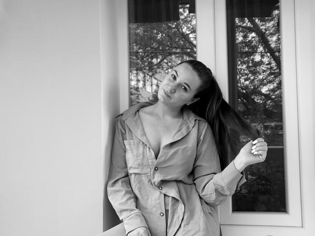 HotAngelsky with long ponytail, wearing unbuttoned button-up shirt, showing cleavage, leaning against window, holding hair, smiling, black-and-white photo.