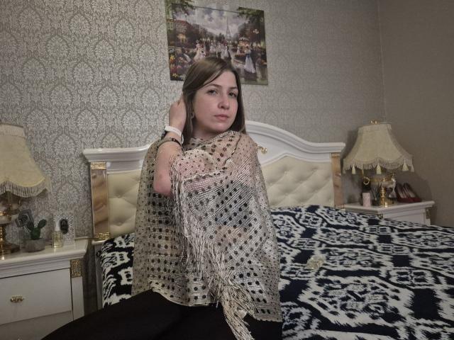 LoriEla with brown hair, wearing a sheer, patterned shawl, black pants, sitting on a bed with a blue and white patterned blanket, white headboard, and two lamps.
