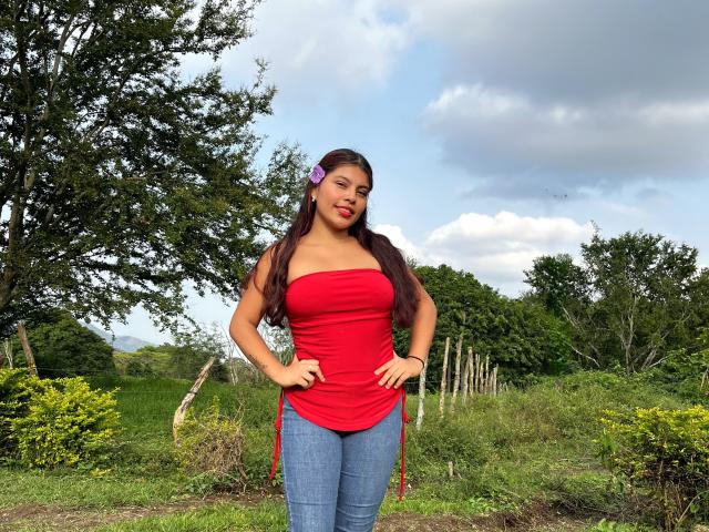 AlyssaGreen stands in a grassy outdoor setting with a cloudy sky. She has long, dark brown hair with a purple flower clip on the left side. She's wearing a red strapless top that shows her shoulders and blue jeans. Her hands are on her hips, and she looks directly at the camera with a slight smile. The greenery and trees in the background contrast with her bright red top. Her skin is a medium brown tone. She's wearing a black bracelet on her right wrist.