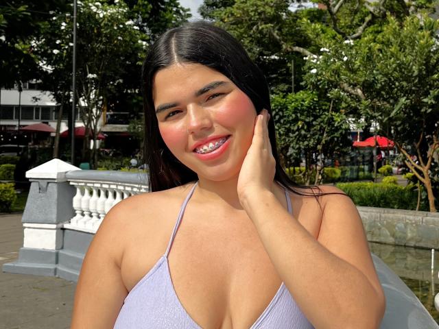 MiluJones, a cute Latina woman with dark hair, pink blush on cheeks, and a purple halter top, smiling with hand on cheek, showing her tits. Sunlit outdoor setting.