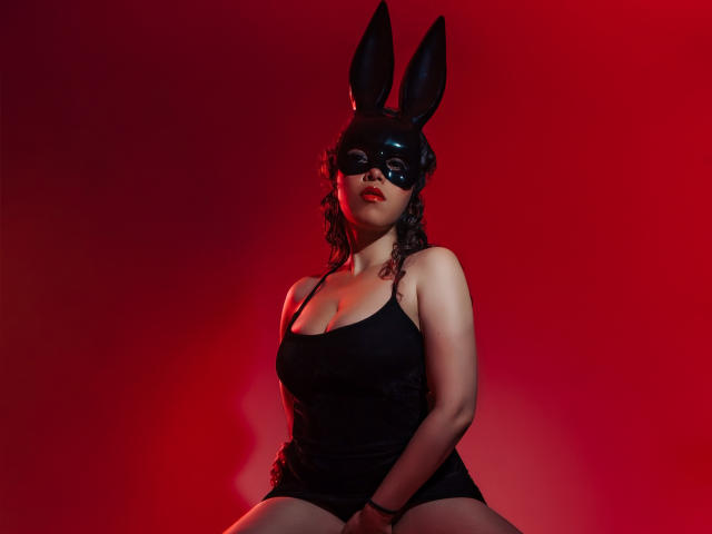 BarbaraLeythonn with black bunny mask, red lipstick, curly hair, black tank top, showing her tits, seated against red light, hands on knees, looking at camera.