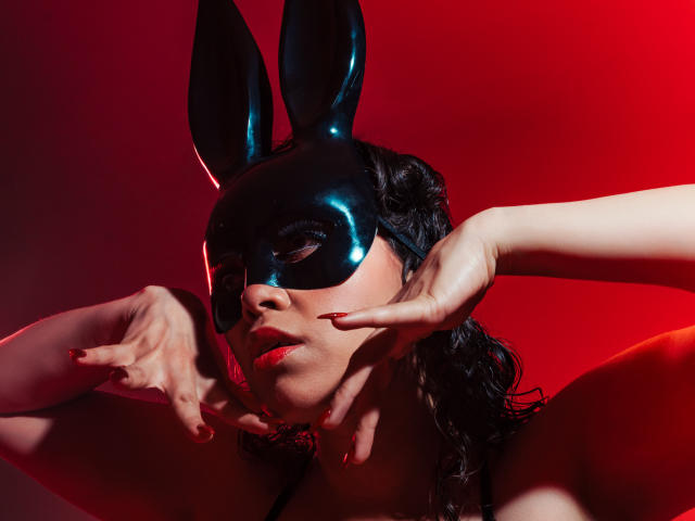 Barbara Leythonn, avec un masque de lapin noir, des ongles rouges et des cheveux noirs ondulés, touchant son visage, éclairage rouge, masque brillant, regard séduisant, buste, sexy, éclairage sombre..