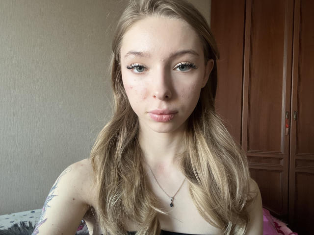 VivanWisseman has light brown wavy hair, green eyes, a small nose, and subtle makeup. She's wearing a necklace with a black pendant, showing a tattoo on her right arm.