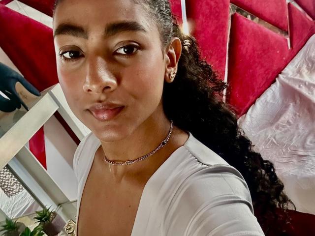 NatalyBlonder has medium brown skin, curly black hair, small gold earrings, and is wearing a white top with a silver choker. She's smiling slightly, looking at the camera.