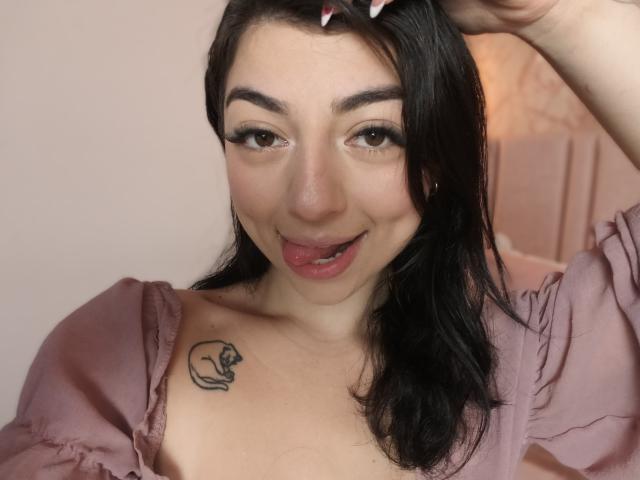 AnntonellaWatson with black hair, pink shirt, and small tattoo on right breast, sticking out tongue, holding hair, brown eyes, smiling, light skin, pink lips, showing nipples.