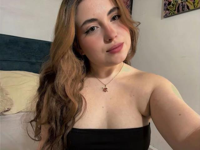 VicluX is a young woman with long, wavy brown hair and fair skin, wearing a strapless black top. She has a subtle smile, light makeup with pinkish lipstick, and a small nose ring. She's wearing a delicate gold necklace with a small red pendant. The background is a bedroom with a dark headboard and a partially visible colorful artwork. The image is a close-up selfie, capturing her from the chest up. Her expression is relaxed and slightly confident.