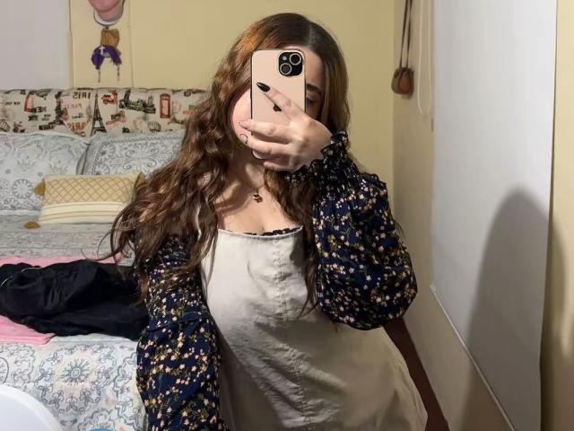 VicluX takes a mirror selfie, holding a pink phone with a black camera. She has long, wavy brown hair covering part of her face. She wears a white dress with a black floral cardigan. The room has a patterned bed with a pink pillow, and a wall with colorful tape and a small bag. The lighting is soft. Her nails are black. The image has a casual, everyday feel.