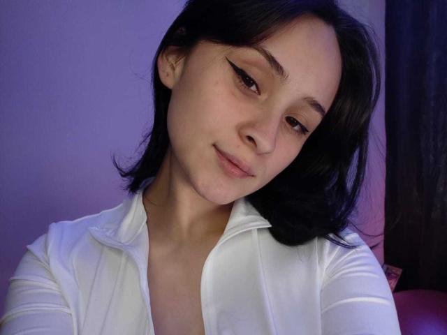 AnnieLamberth has light skin, dark brown shoulder-length hair, and thin black eyeliner. She's looking slightly downward with a subtle smile. She's wearing a white, partially unbuttoned shirt. The lighting is purple-blue, highlighting her face. Her hair is straight and slightly parted to the side. The image has a casual, intimate feel. AnnieLamberth's face is centered, and her head is slightly tilted to the right.