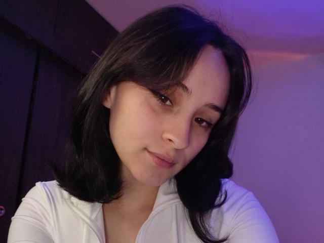 Close-up portrait of AnnieLamberth with a soft, neutral expression. AnnieLamberth has medium-length, straight black hair with subtle bangs. She has light skin, brown eyes, and a small, rounded nose. She wears a white, long-sleeved top. The lighting is purple-tinged, creating a subtle gradient on her face. Her head is slightly tilted to the right. The image has a casual, intimate feel.