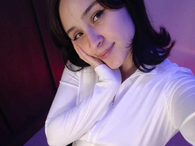 AnnieLamberth takes a selfie, showing a close-up of her face and upper torso. She has medium-length, wavy black hair and is smiling slightly with her right hand resting on her cheek. She wears a white long-sleeve shirt. The image has a soft purple and pink lighting effect, giving it a gentle, almost dreamy atmosphere. AnnieLamberth's skin is fair, and her eyes are slightly downcast, adding a sense of casualness to the photo. The composition is tight, focusing primarily on her face and upper chest.