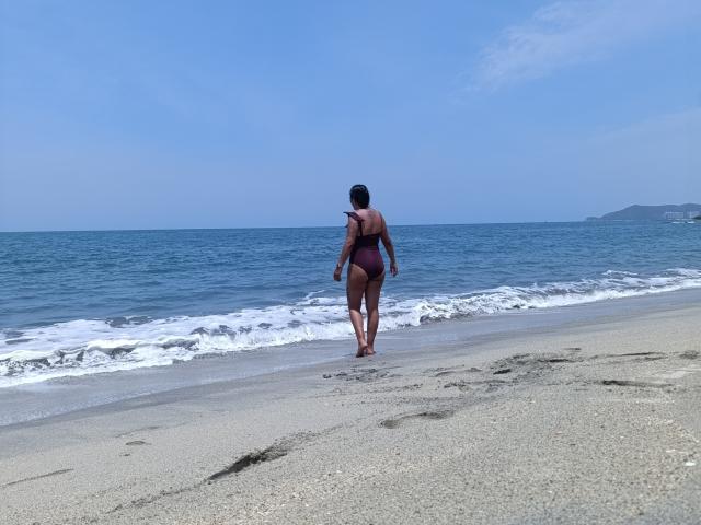A photograph of PauletteLoveX standing on a sandy beach, facing the ocean. She's wearing a dark maroon one-piece swimsuit, with her back to the camera. Her skin is medium brown, and she has short black hair. She's walking towards the water, with small waves gently touching the shore. The sky is clear and blue, and a distant shoreline is visible on the right. Her feet leave subtle imprints in the wet sand. The composition emphasizes the contrast between the beach's texture and the smooth, blue ocean.