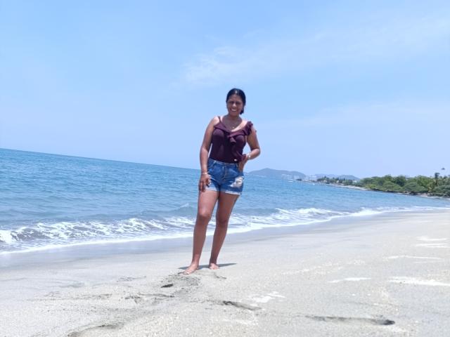 PauletteLoveX stands on a sandy beach with blue ocean waves in the background. She wears a dark purple off-shoulder top and denim shorts. Her dark hair is tied back, and she is barefoot. The clear blue sky contrasts with the green shoreline on the right. She has a medium build and dark skin. Her right hand touches her shorts. The beach has a few footprints and a slightly wet texture near the water.