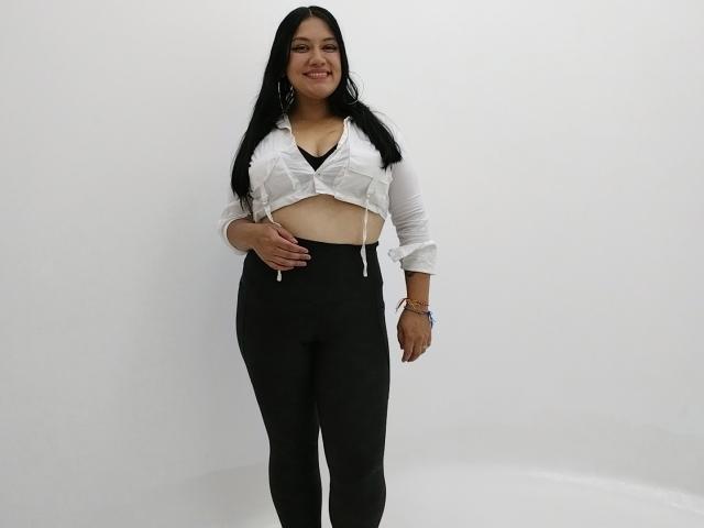 GreyRose with long black hair, white tied-up shirt showing her tits, black high-waisted pants, smiling, standing, wearing bracelets, white wall background.