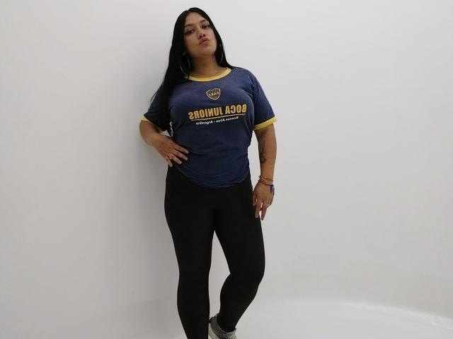 GreyRose with long black hair, wearing a blue Brown Booms t-shirt and black pants, standing against a white wall, hand on hip, looking slightly down, confident.