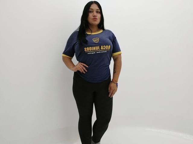 GreyRose stands with hands on hips, wearing a blue "Shovin' Good" shirt, black leggings, and a bracelet, with long black hair and a neutral expression.