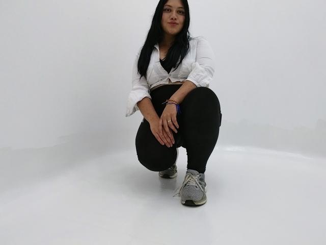 GreyRose with long black hair, crouching, wearing a white tied-up shirt, black pants, and grey sneakers. She has a medium butt, hands on knees, looking at camera.