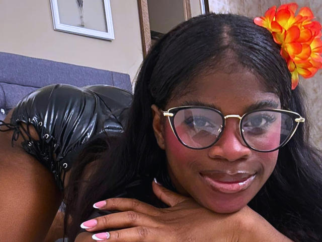 TamaraLubezki has dark skin, long black hair with an orange and yellow flower on the right, and wears gold-framed glasses. She's smiling with pink lipstick, showing white teeth. Her right hand with pink nails rests on a black leather couch. She's wearing black shiny leather shorts. The image is a close-up selfie. Her glasses have a slight reflection. The background includes a framed picture and a door.
