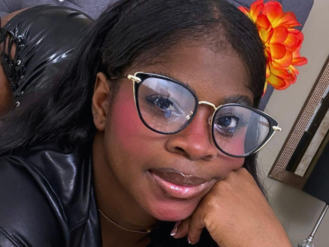 Close-up of TamaraLubezki with dark brown skin, wearing large, gold-framed glasses, and a black leather jacket. Her face is slightly turned to the right, resting her cheek on her hand. She has pink blush on her cheeks, shiny lips, and long black hair with a bright orange flower clip. The background includes a blurred grey figure and a colorful orange flower. The image is a photograph with a casual, candid style.