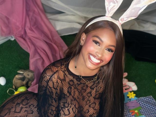 LiaTrainor with brown skin, long brown hair, wearing a white bunny ear headband, black lace dress, smiling, showing her teeth, and wearing a necklace, sitting on green grass.