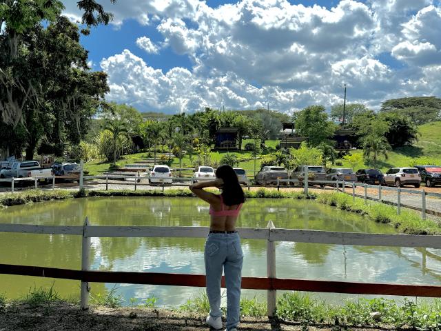 KimmyK stands with her back to the viewer, hands in her hair, looking over a small, greenish pond. She wears a pink bra and blue high-waisted jeans. The white and red wooden fence in the foreground contrasts with the lush greenery and cloudy blue sky. Cars are parked on the other side of the pond. Trees and small buildings are in the background, with a hill rising in the distance. The composition uses natural light and vivid colors.