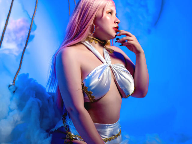 Nyxaria met roze haar, een witte satijnen bikini, gouden kettingen en oorbellen, met een vinger op haar lippen, verlicht met blauw licht, waarbij haar borsten en een sexy profiel zichtbaar zijn..