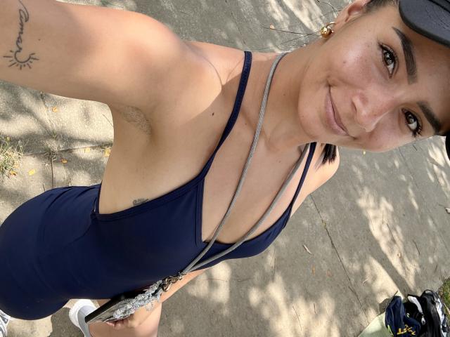 KimmyK, a young woman with light brown skin and dark hair, takes a selfie. She's wearing a navy blue, strappy, one-piece swimsuit that reveals her small, perky breasts and a tattoo on her right arm. She's also wearing a black cap and gold earrings. Her right armpit is slightly visible. She stands on a sunlit, leaf-strewn path with a bag of sports gear in her left hand. The image is sharp, with natural light and shadows.