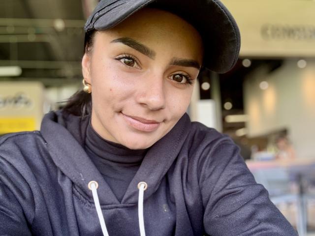 Close-up of KimmyK, a young woman with olive skin and dark eyebrows, wearing a black cap and a navy hoodie. She has brown eyes, a small nose, and a slight smile. Her dark hair is mostly hidden under the cap. She has small gold earrings. The hoodie has white drawstrings. The texture of the hoodie is slightly rough. The background is blurred, with indistinct shapes and colors, focusing attention on KimmyK. The image has a natural, casual feel.