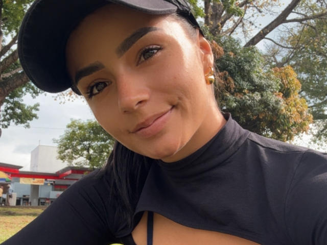 KimmyK with dark hair in a cap, black top, and gold earrings, smiling at the camera, showing a small cut-out in her top, exposing a bit of her tits.