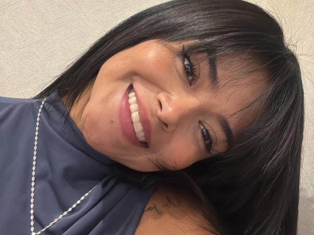 MajoJones has a close-up selfie with a slight tilt. She has straight black hair, dark eyebrows, and is smiling with visible teeth. She wears a navy blue sleeveless top with a high neck and a silver chain necklace. Her skin is smooth and she has a small tattoo on her right shoulder. The lighting is even, highlighting her natural makeup and relaxed expression. The texture of the wall in the background is subtly patterned.