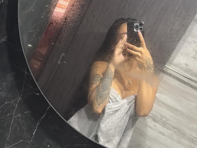 MajoJones with tattoos, taking a mirror selfie, covering her tits with a white towel, wet from the shower, holding a phone in her right hand, water droplets on the mirror.