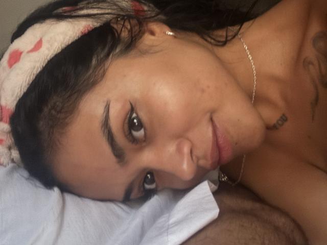 Close-up of MajoJones' face, showing dark, slightly wavy hair with a pink and white headband. Her brown eyes are slightly upturned, with thick, well-defined eyebrows. She has a small tattoo near her right jawline. She's lying on a white pillow, with her head resting on it, and she's smiling softly. Her skin is light brown, and she's wearing a small gold earring. Her lips are slightly parted, and she has a small nose ring in her left nostril. A silver necklace is visible. The image has a natural, candid feel.